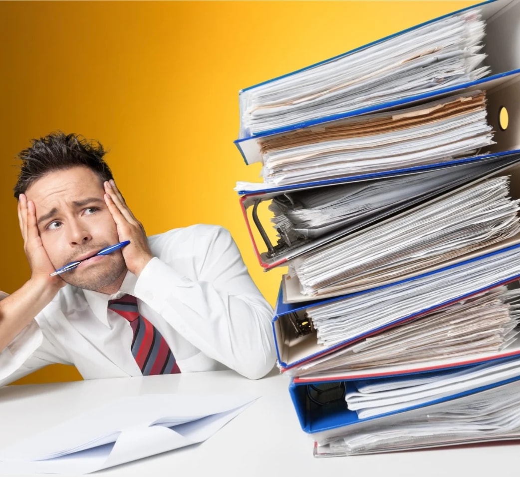 A man at a desk with his head in his hands beside stacks of ring binders.