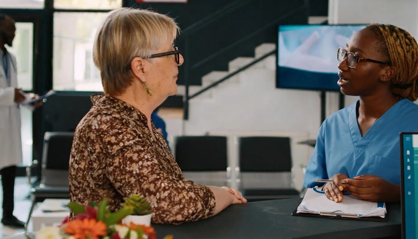 An older woman in conversation with a nurse across a reception desk.