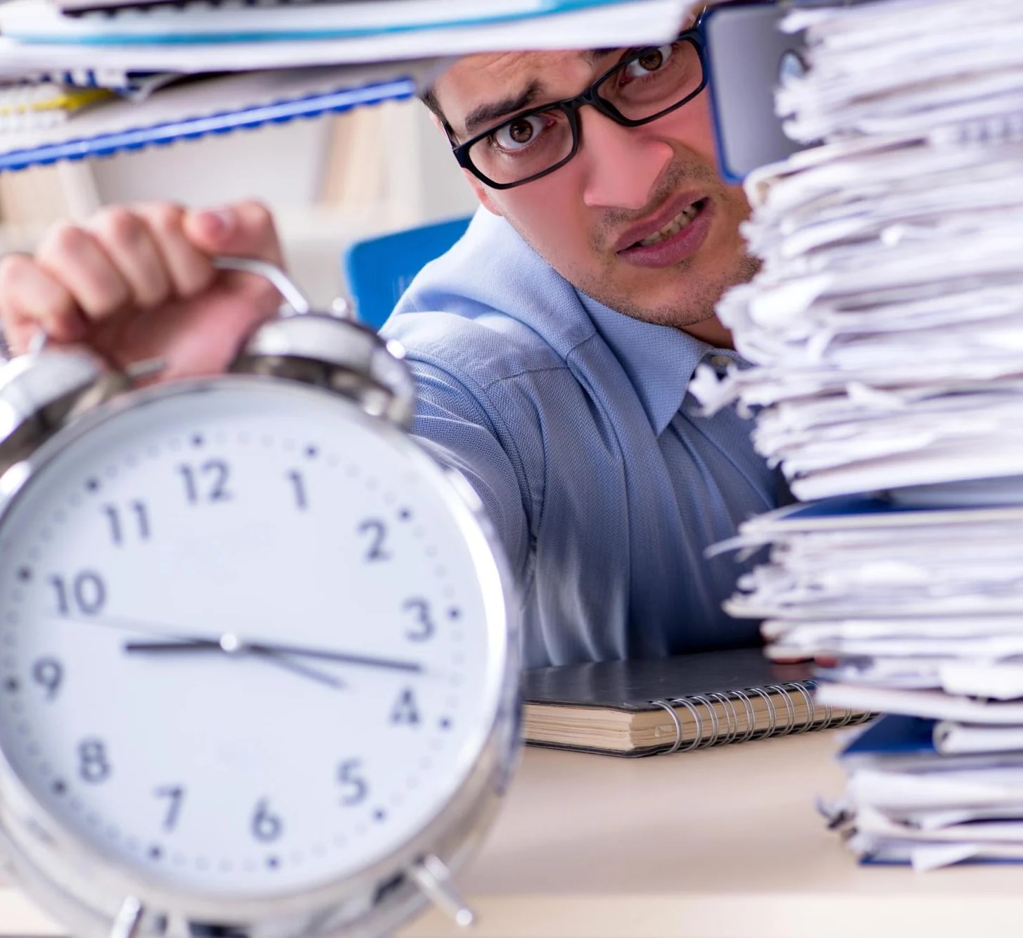 A man peering round a towering stack of paperwork beside an alarm clock.