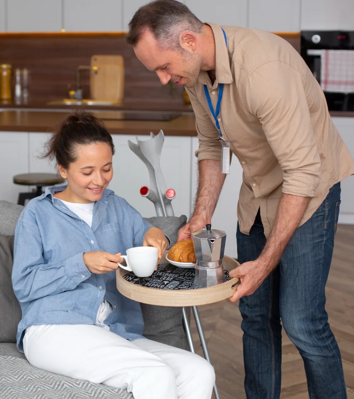 A man bringing coffee and pastries to a woman at a kitchen table.