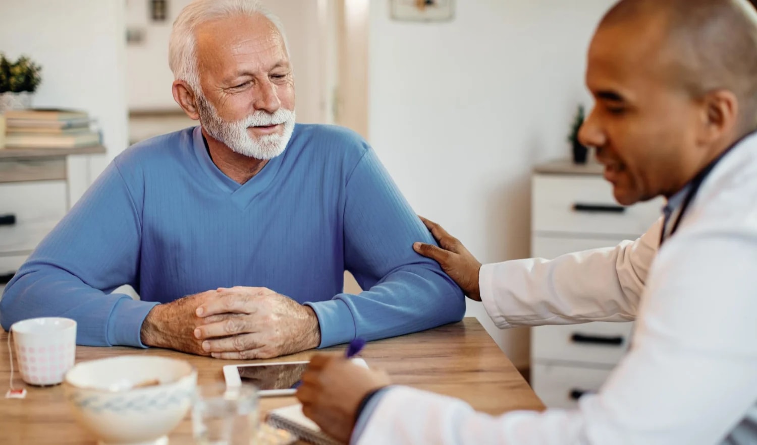 A doctor resting a hand on an older man's shoulder at his kitchen table.