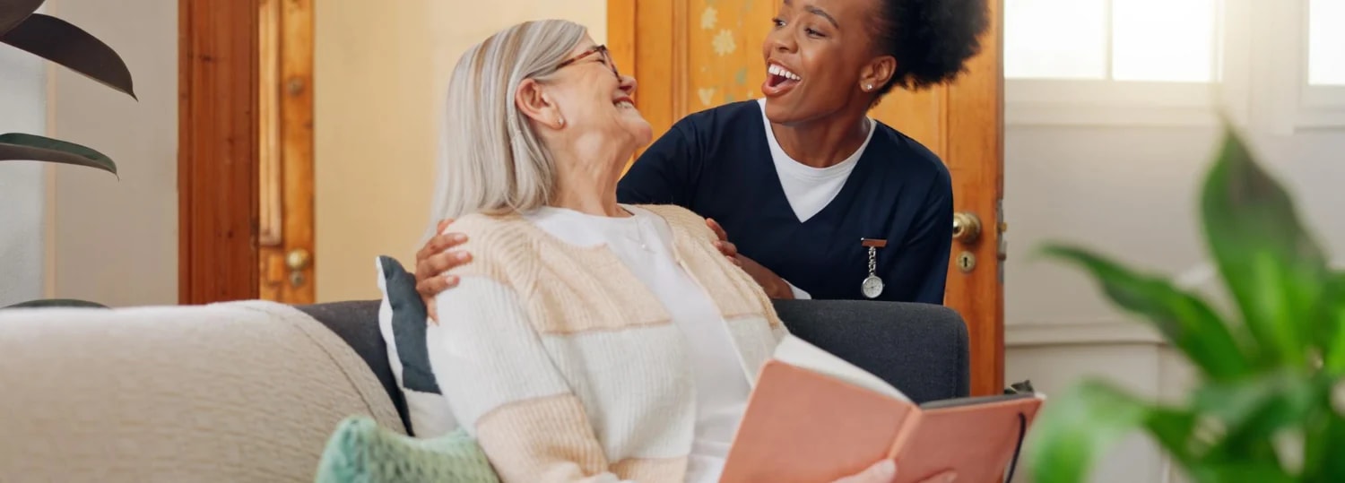An older woman laughing with her carer on the sofa, a book open in her lap.