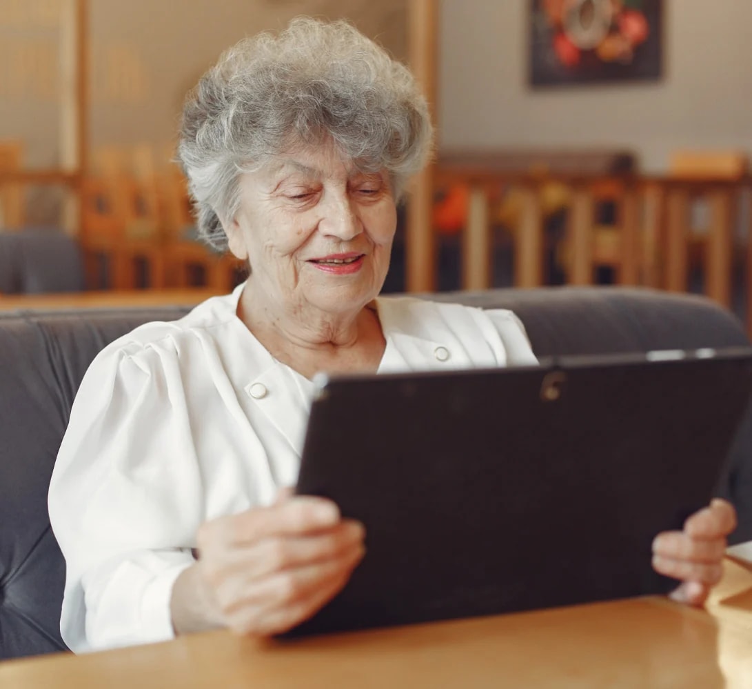An older woman smiling at a tablet propped on the table in front of her.