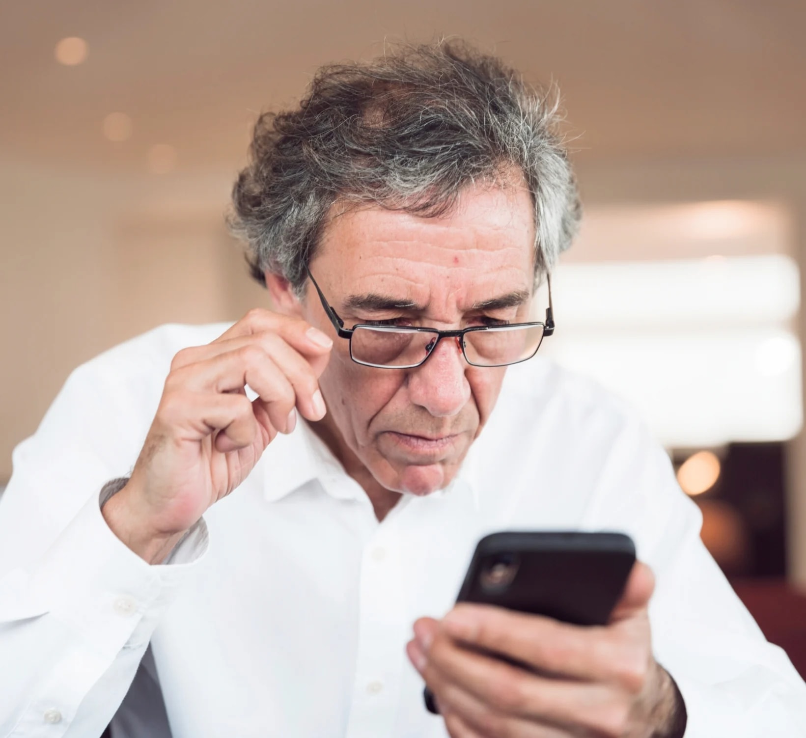 An older man lifting his glasses to read a message on his phone.
